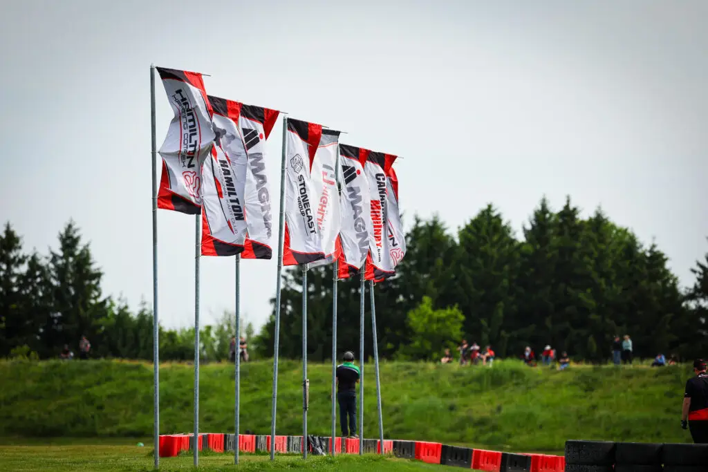 Row of red, white, and black flags on tall poles along a race course, with spectators and trees in the background.
