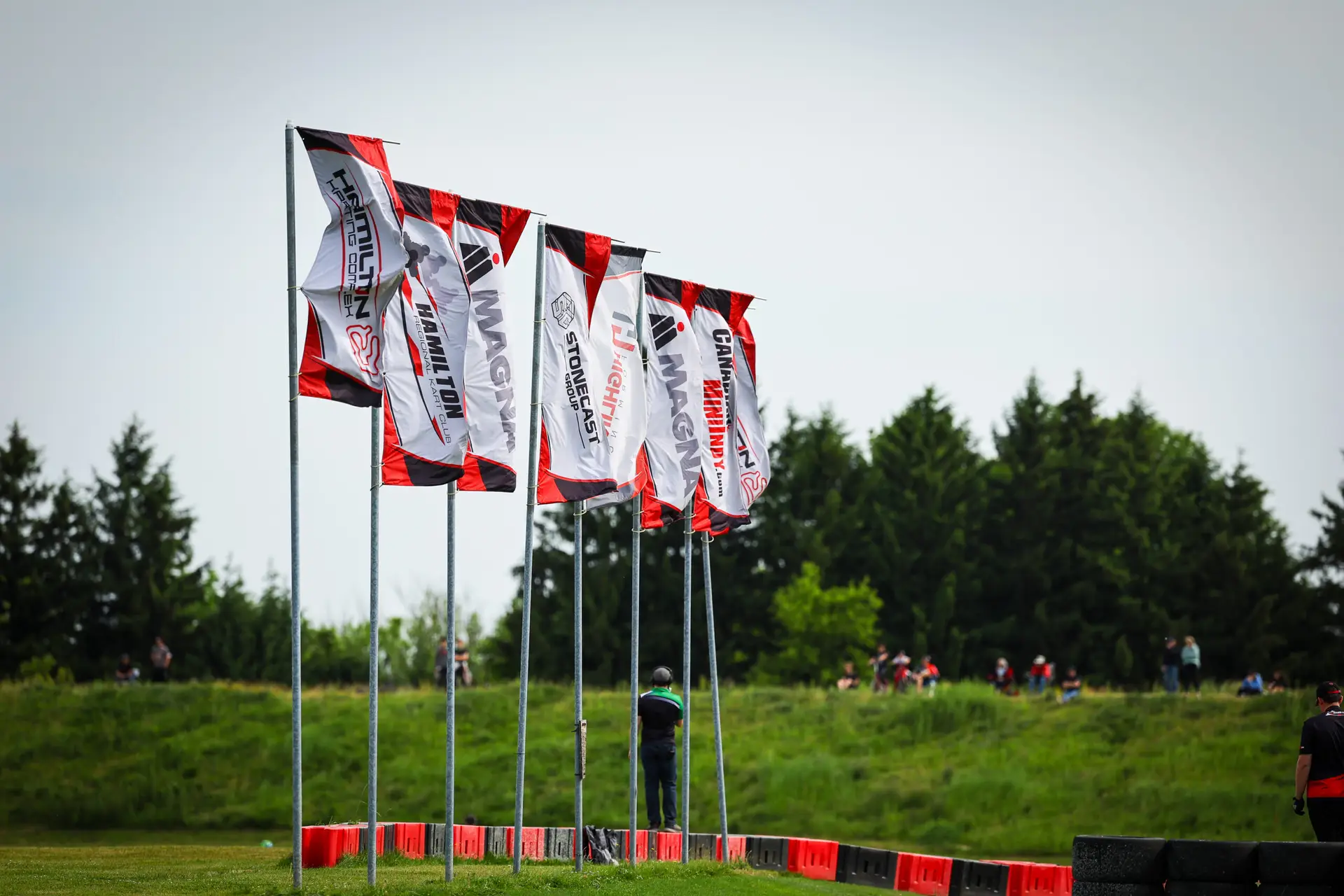Row of red, white, and black flags on tall poles along a race course, with spectators and trees in the background.