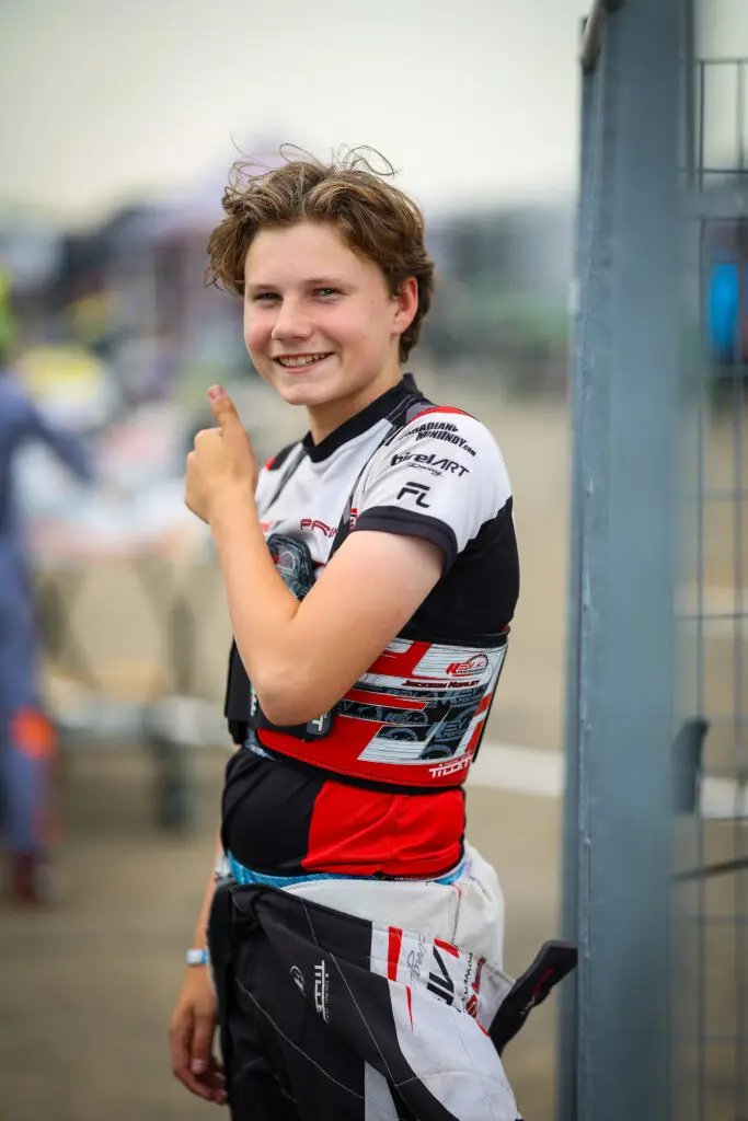 Teenage cyclist in a red, black, and white racing suit smiles and gives a thumbs up beside a fence near the track.