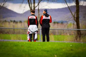 Two people in racing suits stand with their backs to the camera, looking over a chain-link fence at a scenic valley and mountains.