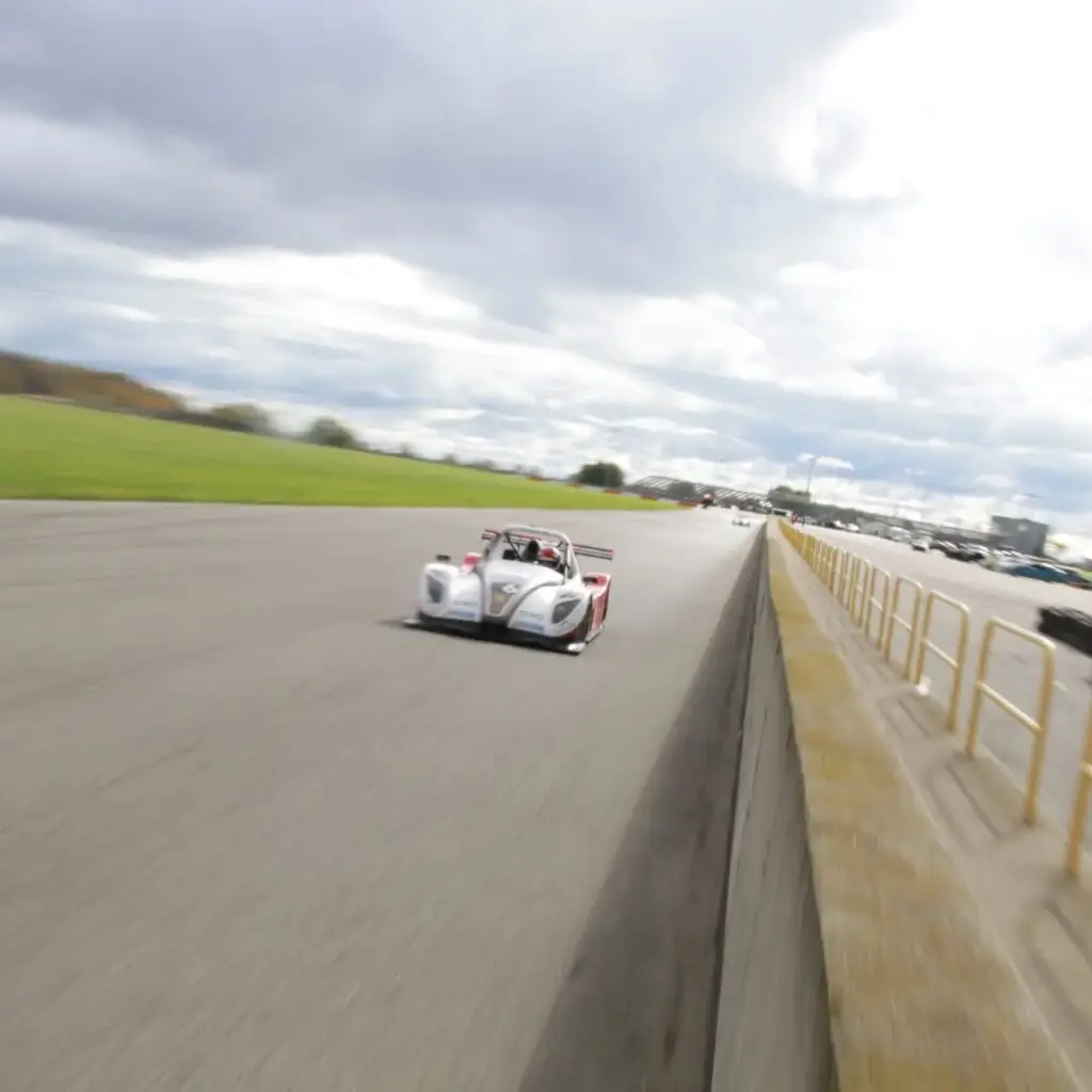 Open-cockpit race car speeding along a track with motion blur and barriers on the right under an overcast sky.