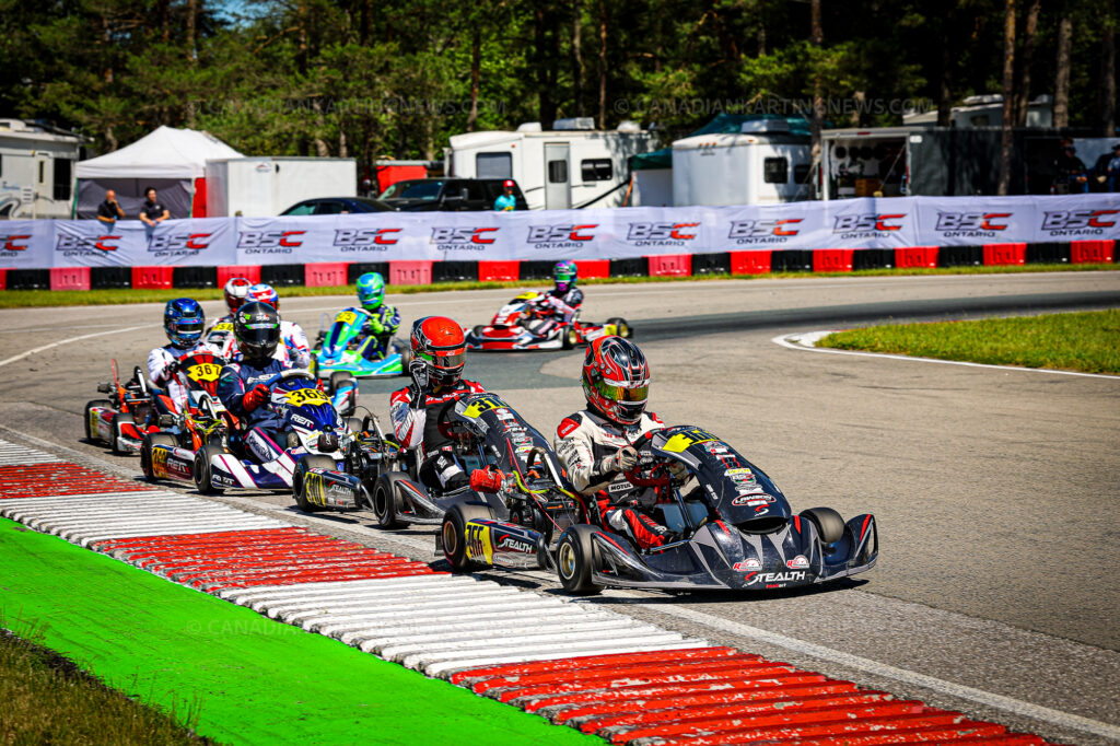 Group of go-kart racers round a bend on a paved track, wearing colorful helmets and racing suits, with red-and-white barriers and banners in the background.
