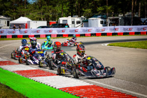 Group of go-kart racers round a bend on a paved track, wearing colorful helmets and racing suits, with red-and-white barriers and banners in the background.