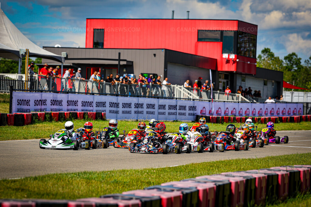 Group of go-karts launching off the start line on an outdoor track, with spectators behind safety barriers and a red modern building in the background.